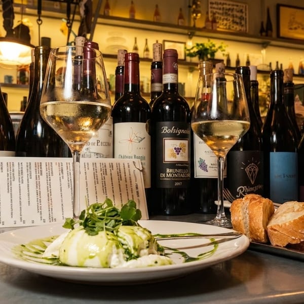 Wine bottles, two glasses of white wine, burrata with pesto, and sliced bread on a bar.