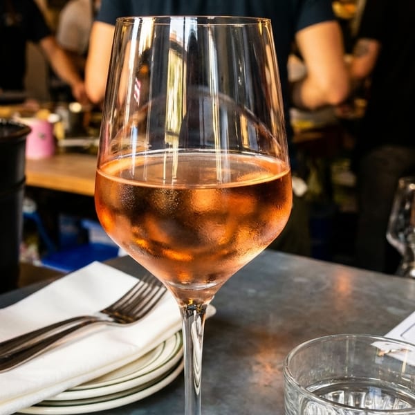 A glass of rosé wine on a table with a menu, plates, and cutlery in a restaurant.