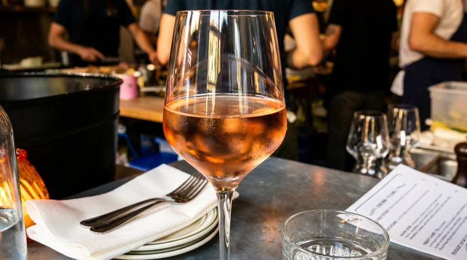 A glass of rosé wine on a table with a menu, plates, and cutlery in a restaurant.